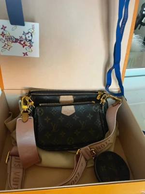 Louis Vuitton Multi Pochette Accessoires - Säljer en Louis Vuitton Multi Pochette Accessoires i monogram canvas med guldiga detaljer. Väskan har flera avtagbara delar, justerbar axelrem i rosa textil och en liten rund myntväska. Klassisk brun färg med LV-logga och beige detaljer. Dustbag, kartong och kvitto medföljer 