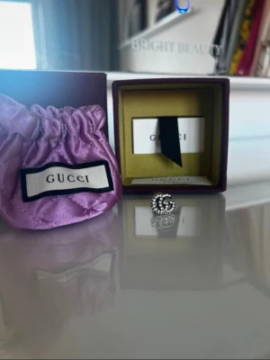 Gucci brosch med strass GG-logo - Snygg brosch från Gucci i form av deras ikoniska GG-logo, dekorerad med glittrande strass. Broschen är i silverfärgad metall och levereras med originalask och lila sammetsfodral. Perfekt accessoar för att lyfta din outfit.