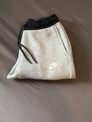 Gråa mjukisbyxor från Nike - Snygga grå mjukisbyxor från Nike med svarta detaljer och dragsko i midjan. Byxorna har sidofickor och Nike-logga tryckt på låret. Perfekta för chill eller träning, tillverkade i mjukt och bekvämt material.
