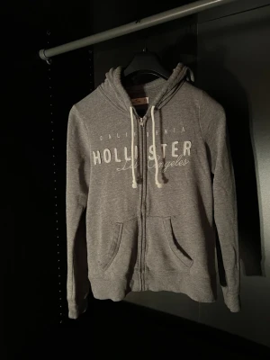 Grå zip-hoodie från Hollister - Grå hoodie med dragkedja från Hollister, med vit logga och texten 'California Los Angeles' på bröstet. Utrustad med huva, vita snören och två stora fickor framtill. Mjuk bomullsblandning och ribbade muddar vid ärmslut och nederkant.