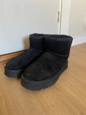 Svarta fluffiga tofflor från Miss Sofie - Mysiga svarta ugg liknande från Miss Sofie med fluffigt foder och grov sula. Skorna är i mocka och har en låg, rundad modell med dekorativ snörning bak. Perfekta för kalla dagar när du vill ha det extra bekvämt och varmt om fötterna.