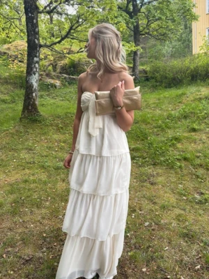 Vit axelbandslös långklänning - Superfin vit långklänning med volanger och axelbandslös design. Krämvit/vit färg. Klänningen har en knytdetalj fram och flera lager i kjolen som ger ett flowy intryck. Perfekt för sommarens tillfällen och har en romantisk vibe. Fint skick! 💕