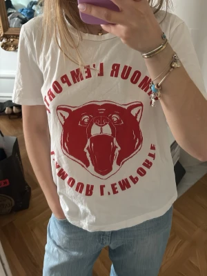 Vit t-shirt med röd björntryck - Vit t-shirt med stort rött tryck av en rytande björn och text på framsidan. T-shirten har en klassisk passform med korta ärmar och rund halsringning. Perfekt för dig som gillar statement-plagg och coola tryck.