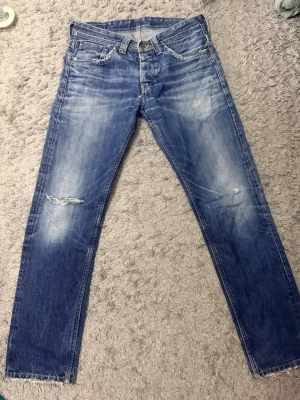 Blå slitna jeans  - Snygga blå jeans med slitningar och ljusa tvättade partier. Klassisk femficksmodell med raka ben och låg midja. Jeansen har en cool, avslappnad look och detaljer som broderad logga på fickan.