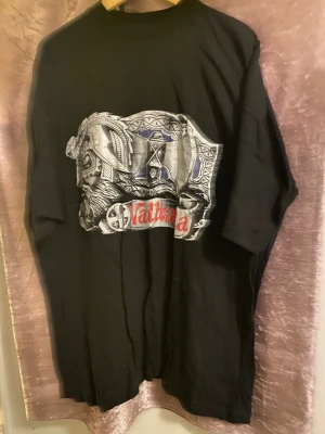 Svart Valhalla t-shirt med tryck - Cool svart t-shirt med stort Valhalla-tryck framtill i silver, blått och rött. Motivet visar en viking med hjälm och detaljerade mönster. T-shirten har rund hals och korta ärmar, perfekt för dig som gillar nordisk mytologi och streetwear. Finns stlk L XL och XXL kvar 120kr/st , helt ny 