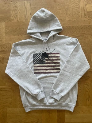 Concrete ink hoodie  - Snygg grå hoodie med huva och dragsko. På bröstet finns ett stort tryck av den amerikanska flaggan med en cool svart färgfläck över stjärnorna. Hoodien har magficka och ribbade muddar. Perfekt för en avslappnad laidback-look. Aldrig använd