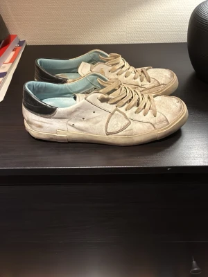 Vita sneakers från Philippe Model - Säljer ett par vita låga sneakers från Philippe Model med svarta detaljer på hälen och ljusblått innerfoder. Skorna har klassisk snörning, klassiska sköldloggan på sidan och är tillverkade i läder. Perfekta för dig som gillar stilrena och trendiga sneakers.