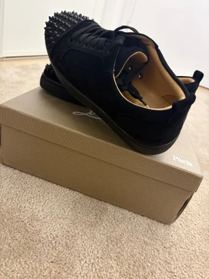 Svarta Louboutin sneakers med nitar - Svarta sneakers från Christian Louboutin med coola svarta nitar på tån och den ikoniska röda sulan. Skorna är låga och har en lyxig känsla med svart mocka och läderdetaljer. Perfekta för dig som vill sticka ut med din stil.