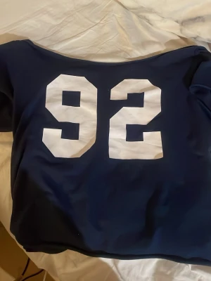 Mörkblå t-shirt med tryck 92 - Snygg mörkblå t-shirt med stort vitt tryck av siffran 92 på ryggen. T-shirten har en klassisk passform och är perfekt för dig som gillar sportig och avslappnad stil. Materialet känns mjukt och bekvämt mot huden.