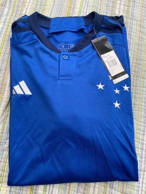 Blå fotbollströja från Adidas - Snygg blå fotbollströja från Adidas med vita detaljer och broderade stjärnor på bröstet. Tröjan har en knapp i halsen och är tillverkad i ett lätt och ventilerande material, perfekt för träning eller match.