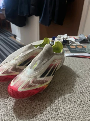 fotbollsskor - Säljer ett par Adidas fotbollsskor i vitt med röda och svarta detaljer samt gula inslag. Skorna har en strömlinjeformad design utan snörning och är tillverkade i syntetmaterial för bästa grepp och känsla på planen. Perfekta för dig som vill sticka ut på matchen.