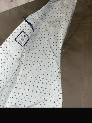 Olivgrön kavaj från Morris - Stilren olivgrön kavaj från Morris med två knappar framtill och tre knappar vid ärmslutet. Kavajen har en vit innerfoder med små svarta mönster och klassisk krage. Perfekt för en dressad look och tillverkad i bomull.