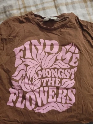 Brun t-shirt med rosa tryck - Brun t-shirt med stort retroinspirerat rosa tryck på framsidan där det står 'Find me amongst the flowers' med blommiga detaljer. Rund halsringning och korta ärmar. Perfekt för en avslappnad och cool stil.