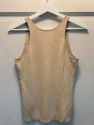 Beige glittrig linne med öppen rygg - Beige ribbad linnetopp med smala axelband och djup öppen rygg. Toppen har en kroppsnära passform och har små glittertrådar i sig som gör att den passar bra på fest
