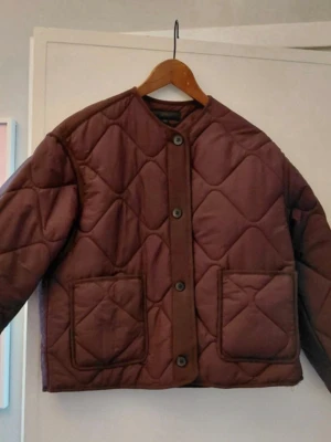Quiltad vinröd vindjacka från Zara - Quiltad vinröd vindjacka från Zara i storlek XS. Jackan har rund hals, stora framfickor och knäppning med knappar framtill. Perfekt för lager-på-lager och har en boxig, lite loose passform. Snygg och enkel att styla till olika outfits.