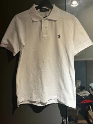 Vit pikétröja från Polo Ralph Lauren - Klassisk vit pikétröja från Polo Ralph Lauren med korta ärmar och krage. Tröjan har knappar framtill och den ikoniska broderade loggan i mörkblått på bröstet. Tillverkad i mjuk bomull för en bekväm känsla.