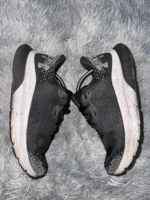 Svarta Under Armour HOVR sneakers - Snygga svarta sneakers från Under Armour med HOVR-sula. Ovandelen är i meshmaterial för bra andningsförmåga och komfort. Skorna har vita och svarta detaljer samt en robust, dämpad sula som passar perfekt till streetwear och träning.