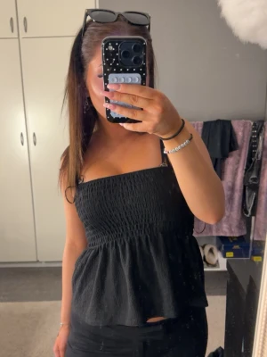 Svart smockad linnetopp - Säljer en svart linnetopp med smockad överdel och tunna axelband. Toppen har en volang nedtill som ger en luftig och söt look. Perfekt till jeans eller kjol för en trendig outfit.