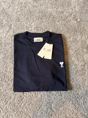 Ami Paris T-shirt mörkblå Ny-oanvänd (M) - AMI Paris T-shirt – Mörkblå (Storlek M) Säljer en helt ny och oanvänd AMI Paris T-shirt i storlek M. Stilren och modern design i mörkblå färg med AMI Paris ikoniska logga som ger en exklusiv och minimalistisk look. Tillverkad i hög kvalitet med mjukt och bekvämt material – perfekt för både vardag och streetwear-stil. 🔹 Märke: AMI Paris 🔹 Modell: T-shirt 🔹 Färg: Mörkblå 🔹 Storlek: M 🔹 Skick: Helt ny / oanvänd 🔹 Passform: Klassisk Nypris är högt –perfekt möjlighet att göra ett fy