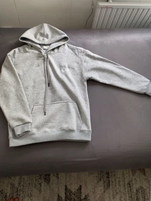Ljusgrå hoodie med ficka - En stilren ljusgrå hoodie med huva och dragsnören. Hoodien har en stor magficka och ribbade muddar vid ärmslut och nederkant. Perfekt för en avslappnad look och enkel att matcha med andra plagg. Den är i storlek M men jag skulle säga den passa S bra 