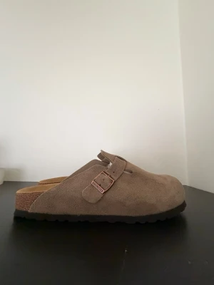 Beige Birkenstock tofflor i mocka - Säljer ett par klassiska beige tofflor från Birkenstock i mjuk mocka. Modellen har en justerbar rem med metallspänne och en bekväm korksula. Yttersulan är i svart gummi med mönstrad struktur för bra grepp. Perfekta för chill dagar hemma eller på stan.