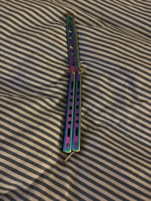 Regnbågsfärgad butterfly-träningskniv - Säljer en butterfly-träningskniv i metall med regnbågsfärgad finish. Handtaget har flera hål och cool design, perfekt för att öva tricks och flips. Kniven är inte vass och passar för dig som vill lära dig hantera butterfly utan risk.