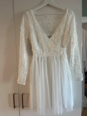 Vit spetsklänning från Bubbleroom - Superfin vit kortklänning från Bubbleroom med v-ringning och långa ärmar. Klänningen har spets med blommigt mönster upptill och en luftig kjol i tyll. Perfekt för sommarens alla tillfällen!