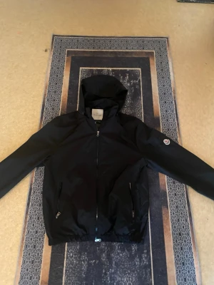 Svart Moncler vindjacka med huva - Snygg svart vindjacka från Moncler med huva och dragkedja framtill. Jackan har två fickor med dragkedja och en Moncler-logga på ärmen. Tillverkad i ett lätt och glansigt material som skyddar mot vind. Perfekt för en stilren och sportig look. Helt ny nästan aldrig använd, säljer den pågrund av att den blev för stor.