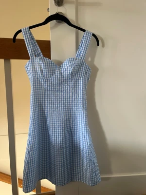 Blåvit rutig sommarklänning - Söt blåvit rutig klänning med breda axelband och markerad byst. Klänningen har smock i ryggen för extra komfort och är i en kort modell. Perfekt för varma dagar och har en klassisk, fräsch look.