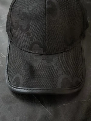 Svart keps  med GG-mönster - Snygg svart keps  med det klassiska GG-monogrammet ton-i-ton över hela kepsen. Tillverkad i bomull och polyester med detaljer i läder. Perfekt accessoar för en trendig streetstyle-look.