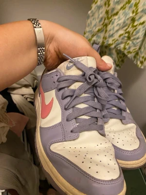 Nike Dunk Low lila och rosa - Nike Dunk Low sneakers i vitt och lila läder med rosa swoosh. Klassisk siluett med perforerad tå, platta lila skosnören och matchande yttersula. Perfekta för dig som vill sticka ut med färg och stil.