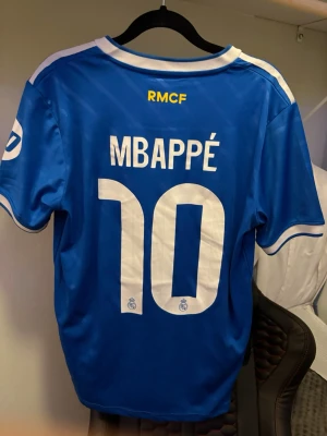 Real Madrid Mbappé matchtröja blå - Snygg blå Real Madrid matchtröja med Mbappé och nummer 10 på ryggen. Tröjan har vita detaljer, klubbmärke och Adidas-logga på bröstet samt sponsortryck. Perfekt för fotbollsfans som vill sticka ut på läktaren eller planen.