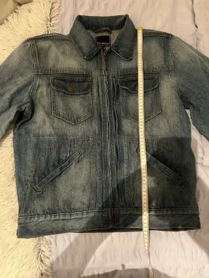 Klassisk blå jeansjacka från Rainbow - Snygg blå jeansjacka från Rainbow med klassisk krage, två bröstfickor med knappar och två snedställda fickor framtill. Jackan har en cool tvättad look och bruna knappdetaljer. Perfekt för dig som gillar en tidlös streetstil.