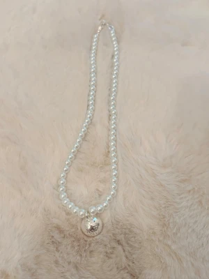 Pärlhalsband från SNÖ med silverberlock - Säljer ett stilrent halsband från SNÖ med vita pärlor och en rund silverfärgad berlock där det står SNÖ. Halsbandet har klassisk design och pärlorna ger en elegant känsla. Perfekt för dig som gillar tidlösa accessoarer.