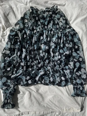 Blommig blus från Bik Bok - Snygg blommig blus från Bik Bok i svart, vitt och ljusblått. Blusen har långärmade puffärmar med smock vid ärmslut och midja, samt volangdetaljer vid halsen. Tunt, lätt material som ger en luftig känsla. Pris kan diskuteras