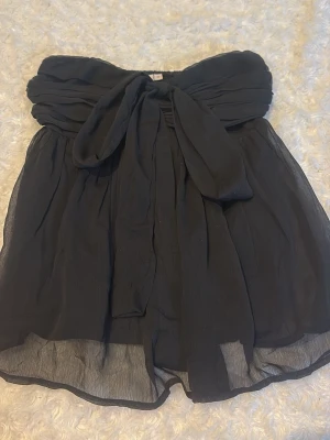 Svart axelbandslös tröja rosett - Svart bandeau tubtopp i luftig syntetmaterial med stor rosett framtill och smockad rygg. Toppen är axelbandslös och har ett flowy, voluminöst fall som ger en trendig och chic vibe. Inte använd💘