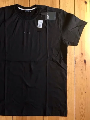 Svart BOSS t-shirt i bomull - Stilren svart t-shirt från BOSS med diskret logga på bröstet. Klassisk rund halsringning och korta ärmar. Tillverkad i mjuk bomull som känns skön mot huden. Perfekt för en clean och enkel look.