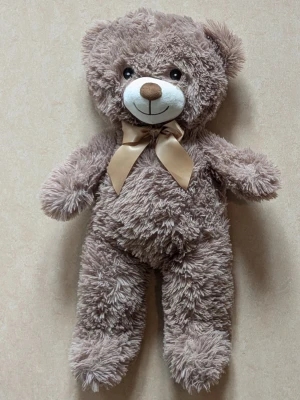 Mjuk nallebjörn med rosett - Säljer en gosig brun nallebjörn med fluffig päls och en ljusbeige rosett runt halsen. Nallen har ett sött broderat ansikte med svartvita ögon och brun nos. Perfekt som present eller mysig inredningsdetalj. Längd ca 57cm