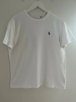Vit t-shirt från Ralph Lauren - Vit t-shirt från Ralph Lauren med blå broderad logga på bröstet. Tjockare material, väldigt bra kvalitet.