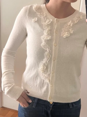 Vit/beige kofta med blommor från Hollister - Supersöt beige/vit kofta från Hollister med dekorativa blommor i tyll längs knäppningen. Koftan har rund halsringning, långa ärmar och pärlemorknappar. Perfekt för dig som gillar romantisk och feminin stil.