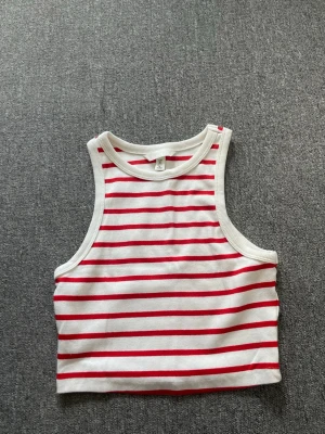 Randig vit och röd linnetopp H&M - Snygg croppad linnetopp från H&M i vitt med röda horisontella ränder. Toppen är ärmlös med breda axelband och rund halsringning. Perfekt för varma dagar och enkel att matcha med jeans eller shorts.