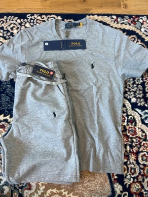 Grått set från Polo Ralph Lauren - Snyggt grått set från Polo Ralph Lauren med t-shirt och shorts. Båda plaggen har den klassiska broderade loggan på bröstet och benet. Setet är gjort i mjukt bomullsmaterial och har en avslappnad passform. Perfekt för en chill dag hemma eller som snyggt lounge-set.