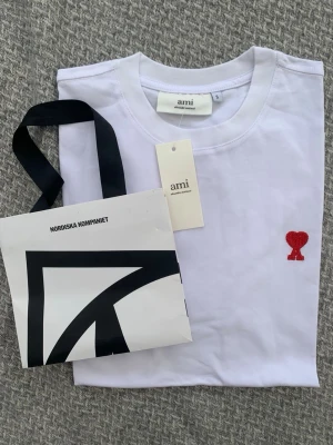 Vit t-shirt från Ami med rött hjärta - Snygg vit t-shirt från Ami Alexandre Mattiussi med ett broderat rött hjärta på bröstet. Klassisk passform och rund halsringning. Tillverkad i mjuk bomull, perfekt för en clean och stilren look.