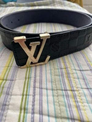  svart rutigt bälte - Snyggt svart bälte från  med klassiskt rutigt mönster och en stor  i guld som spänne. Bältet är gjort i skinn och har en exklusiv känsla som lyfter vilken outfit som helst.