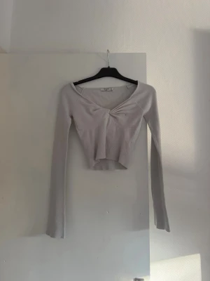 Ljusbeige ribbad topp från NA-KD - Trendig ljusbeige ribbad topp från NA-KD med lång ärm och twistad detalj framtill. Toppen har en croppad passform och v-ringad halsringning som ger en snygg siluett. Perfekt att styla med höga jeans eller kjol. Har sminkfläck