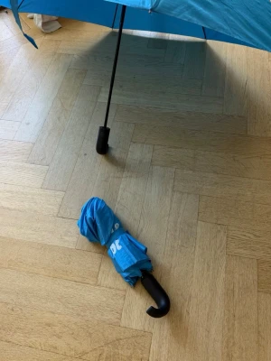 Blått paraply med böjt handtag - Säljer ett stilrent blått paraply med klassiskt böjt svart handtag. Paraplyet är kompakt och lätt att ta med sig, perfekt för regniga dagar. Materialet är syntetiskt och tål vatten bra. Enkel att fälla upp och ihop.