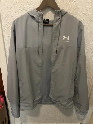 Grå vindjacka från Under Armour(M) Helt ny - Snygg grå vindjacka från Under Armour med huva och dragsko. Jackan har dragkedja framtill, två sidofickor och meshfoder för extra ventilation. Perfekt för blåsiga dagar och sportiga outfits. Stort vitt logotryck på bröstet ger en clean look.