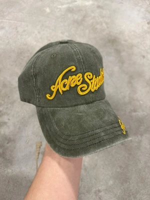 Olivgrön keps från Acne Studios - Snygg olivgrön keps från Acne Studios med gul broderad logga både framtill och på skärmen. Klassisk böjd skärm och justerbar baksida. Tillverkad i mjukt bomullstyg som ger en avslappnad vibe.
