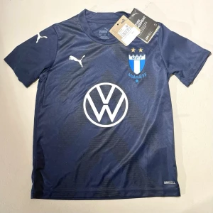 Blå Malmö FF matchtröja Puma - Malmö FF matchtröja för barn i mörkblått , två guldstjärnor och VW-logga på bröstet. Tröjan är tillverkad i syntetmaterial med DryCell-teknologi som håller dig torr under träning. Rund hals och korta ärmar, perfekt för fotboll och sport. Passform är herr men funkar såklart till dam då det är en tröja för barn😄