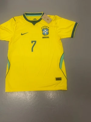 Brasiliens vm tröja Vini - Gul Brasilien fotbollströja från Nike med gröna detaljer, nummer 7 och Vini Jr. på ryggen. Tröjan har korta ärmar, rund hals och officiellt landslagsmärke på bröstet. Perfekt för dig som älskar fotboll och vill stötta Brasilien.